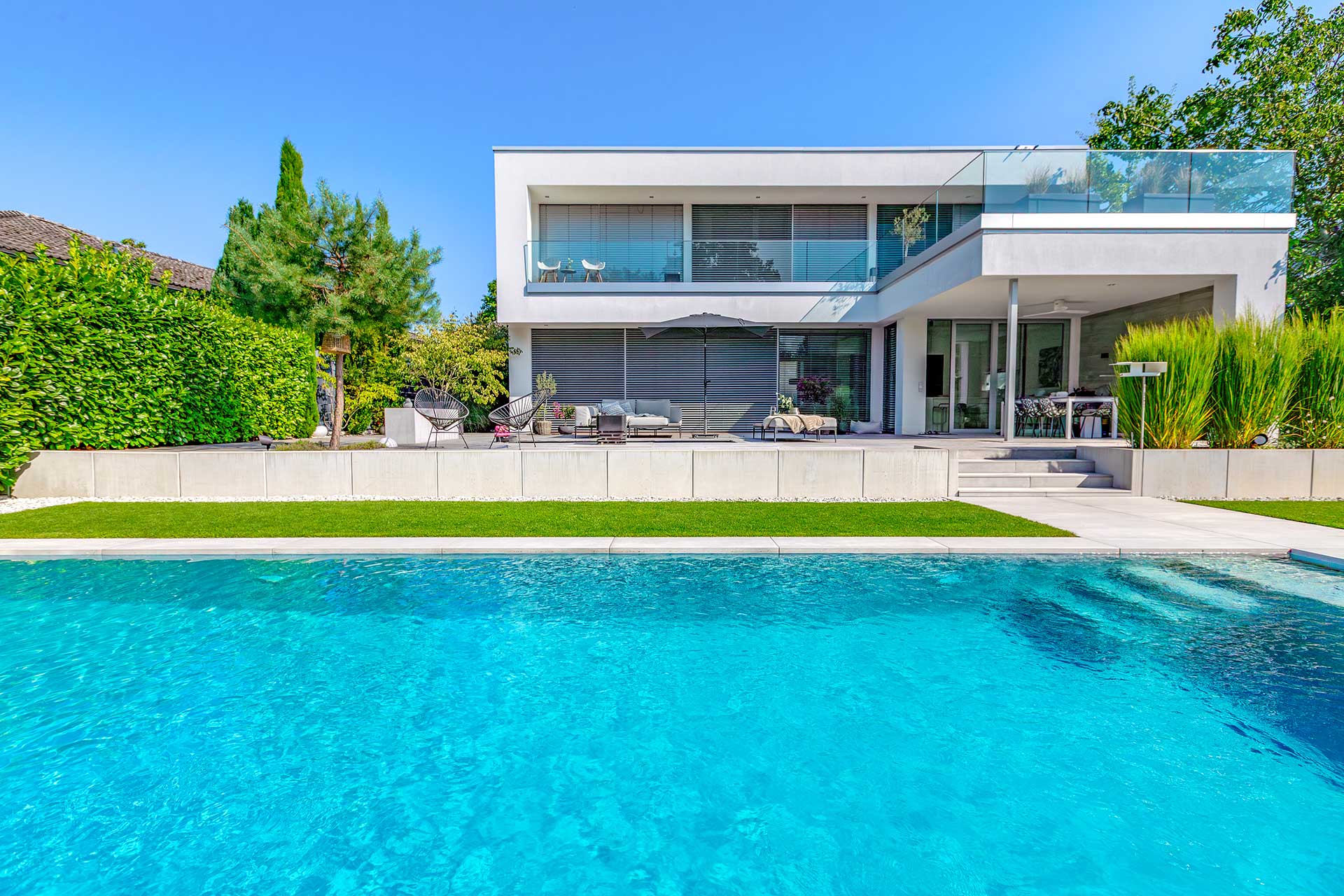 Modernes, weißes Haus mit Glasbalkon und großem Pool im sonnigen Garten