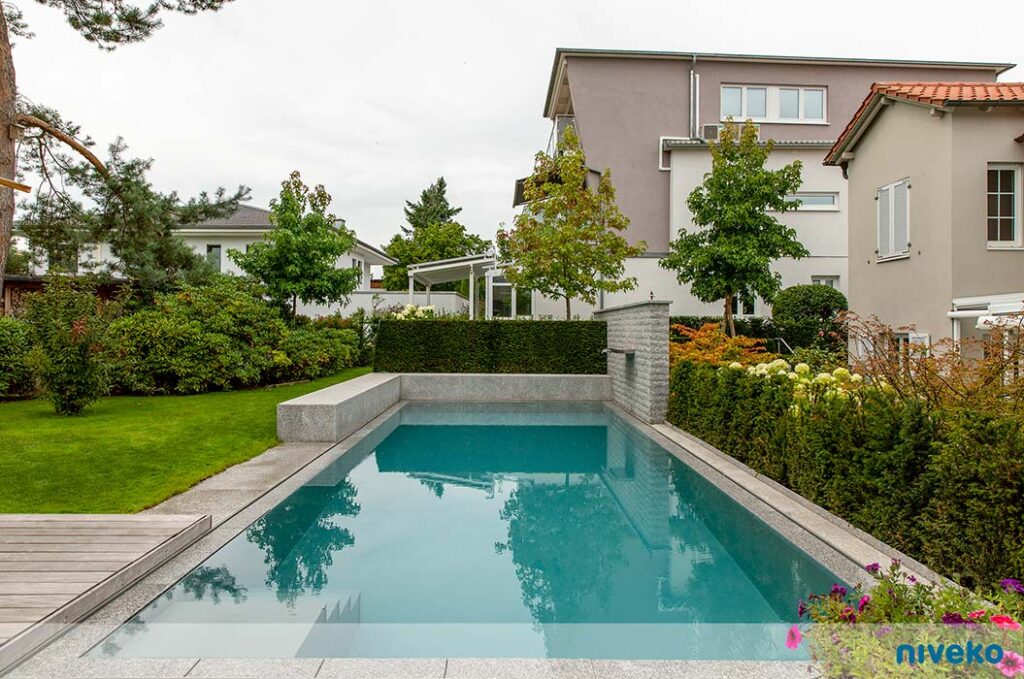 Ein moderner rechteckiger Pool mit grauem Rand spiegelt grüne Sträucher und ein Wohnhaus