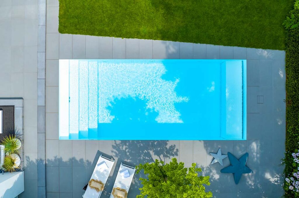 Ein rechteckiger blauer Pool ist von grauen Fliesen und grünem Rasen umgeben