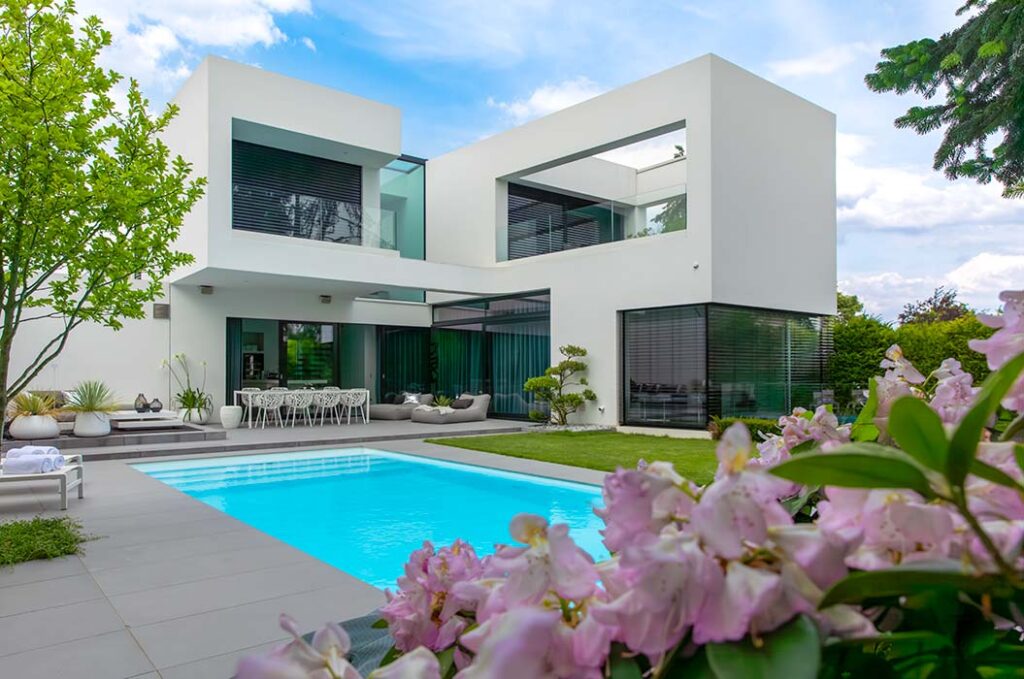 Modernes weißes Haus mit großen Fenstern, Pool und blühenden Pflanzen im Garten