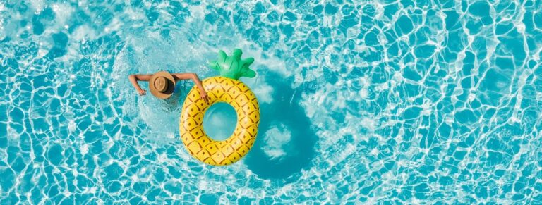 Draufsicht auf eine junge Frau im Bikini und mit Strohhut vor dem Hintergrund blauer Wellen in einem Swimmingpool, mit einer großen aufblasbaren gelben Ananas-Schwimmhilfe. Konzept für entspannte Sommerferien in einem Luxusresort.