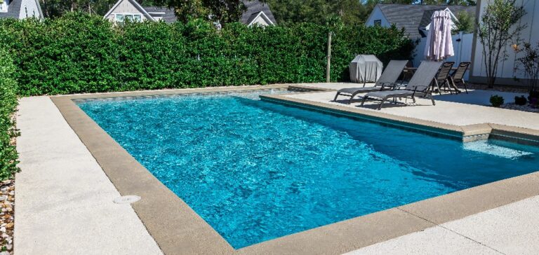 Ein rechteckiger neuer Swimmingpool mit hellbraunen Betonrändern im umzäunten Hinterhof eines Neubaus
