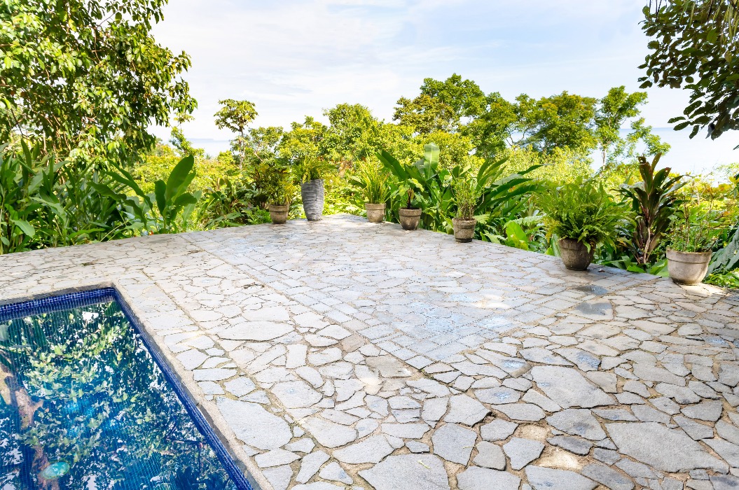 Eine offene und freie Terrasse aus beigem Naturstein neben einem blauen Pool, umgeben von grünen Pflanzen aus dem Regenwald.
