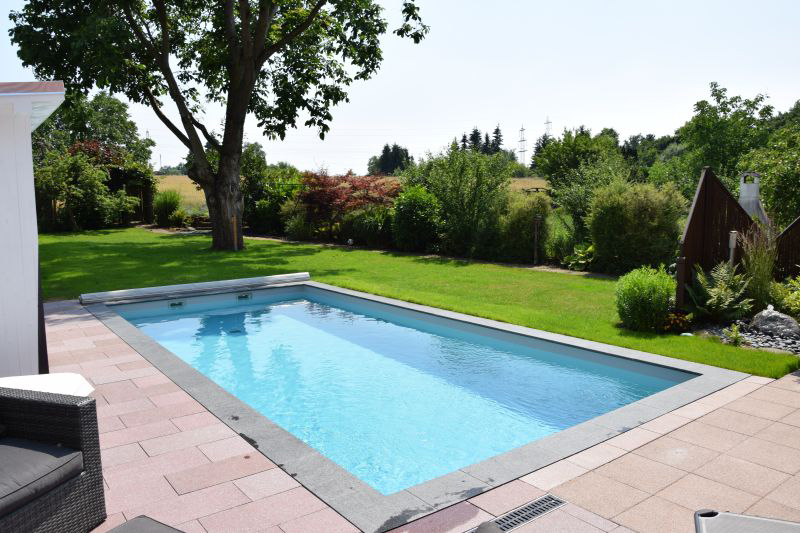Ein moderner Pool im Garten mit klarem Wasser und gruenem Rasen umgeben von Baeumen