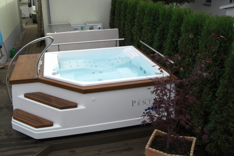 Weisser Whirlpool mit Holzverkleidung und klar blauem Wasser steht neben grüner Hecke