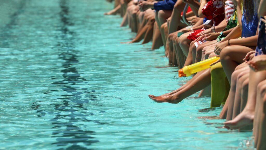 Ein blaues Schwimmbecken, an dessen Rand Schüler sitzen, die ihre Füße und Zehen ins Wasser baumeln lassen und zuschauen. 
